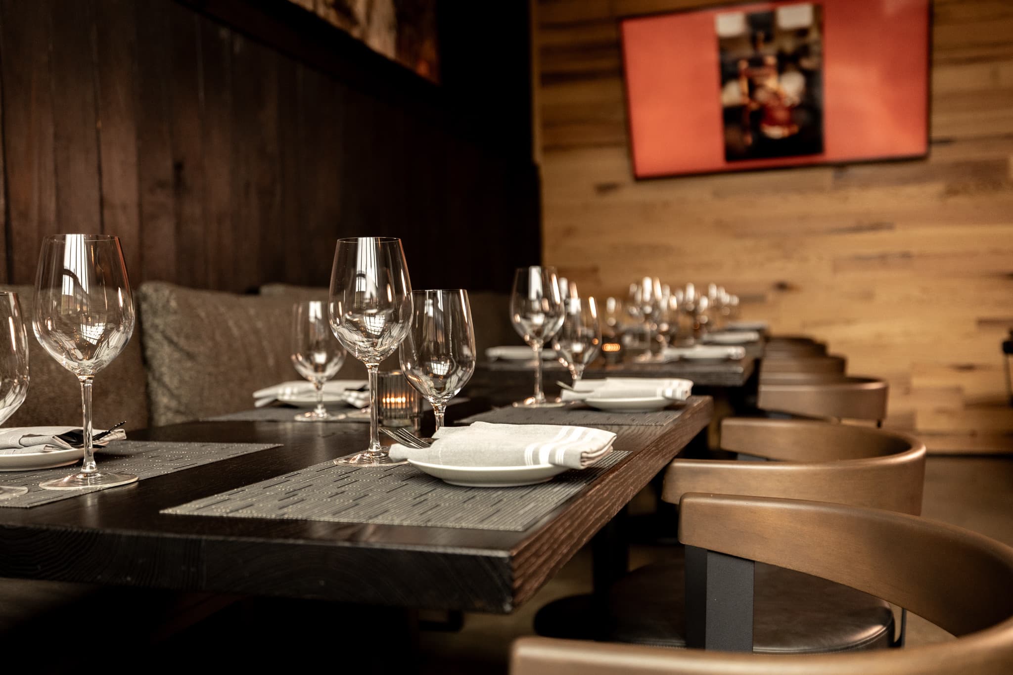 A long table set with wine glasses and white napkins beside the wall-mounted screen in the private dining room at CHR Steak