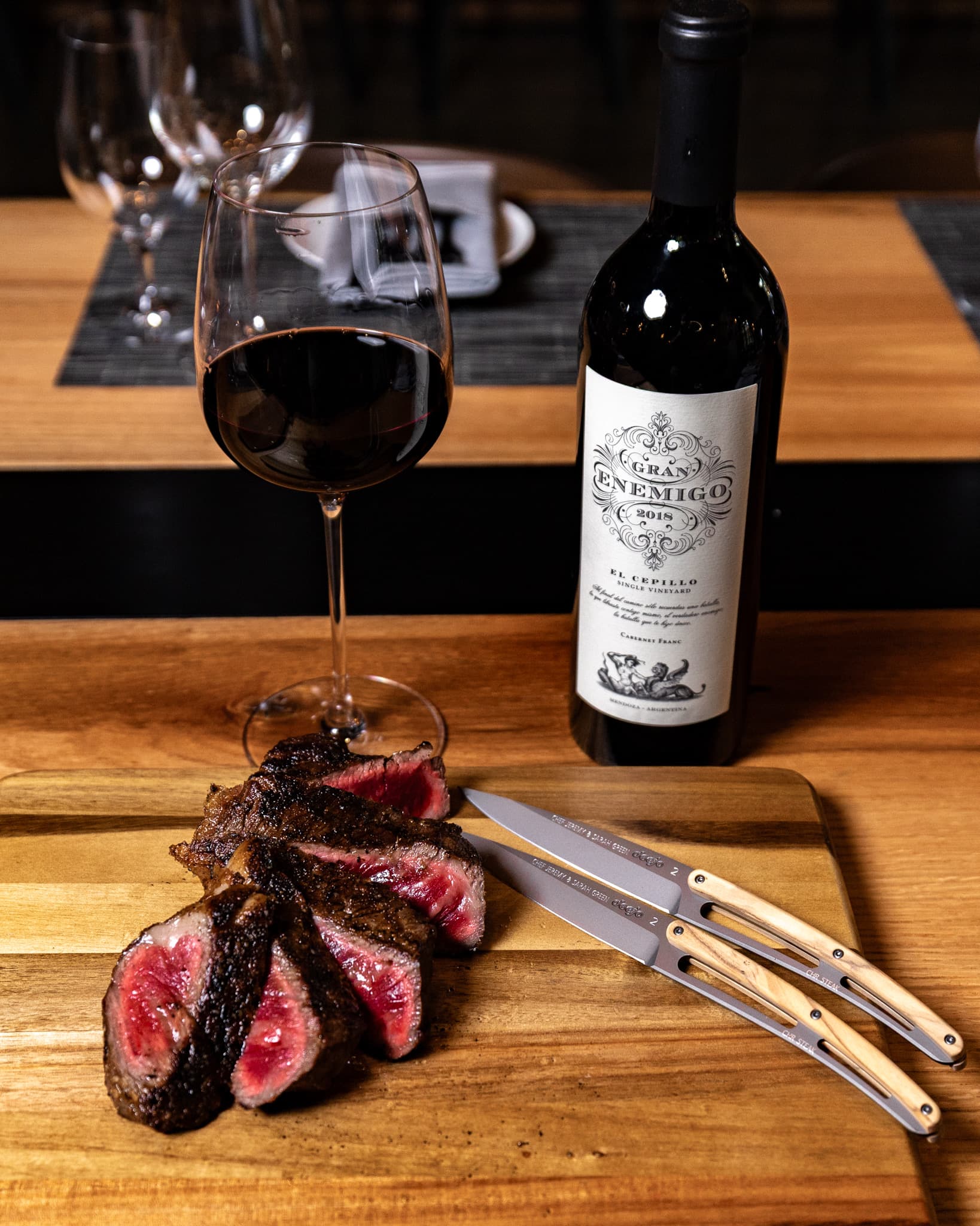 A pair of engraved Blade Club steak knives with olive wood handles resting on a wooden board next to sliced steak at CHR Steak