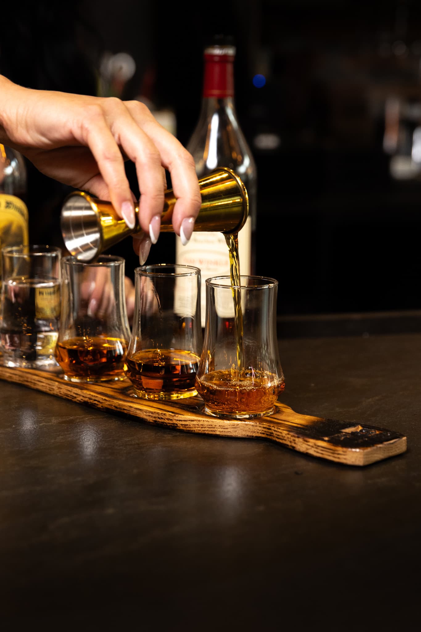 A bartender pouring bourbon from a brass jigger into a tasting glass on a charred barrel stave flight board at CHR Steak