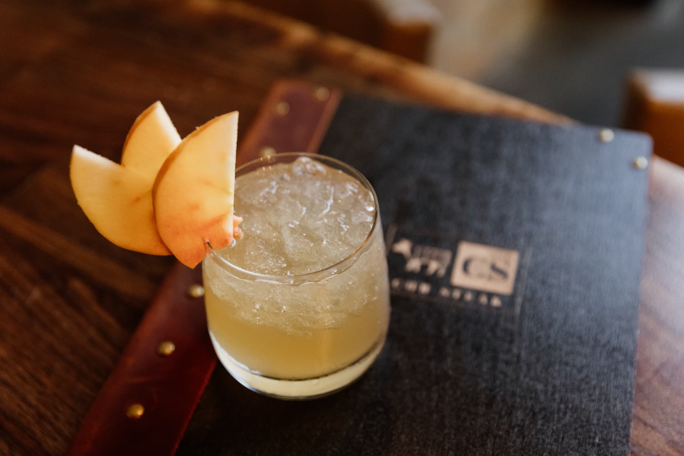 A cocktail over crushed ice garnished with apple slices, beside a CHR Steak menu book